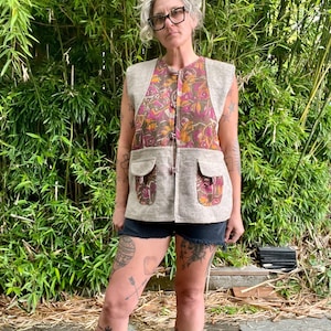 May include: A sleeveless vest with a beige base and a patterned front panel featuring pink, orange, and brown floral designs. The vest has two front pockets and a button closure. The person is wearing black shorts.