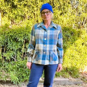 May include: A blue and white plaid shirt jacket with a button front closure and two patch pockets. The jacket is worn over a pair of dark blue jeans and a blue beanie.