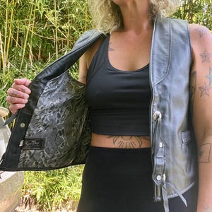 May include: A black leather vest with silver-toned buttons and decorative leather ties. The vest's interior features a patterned lining. The vest is open, revealing a black top and black leggings. The vest is a classic piece of apparel.