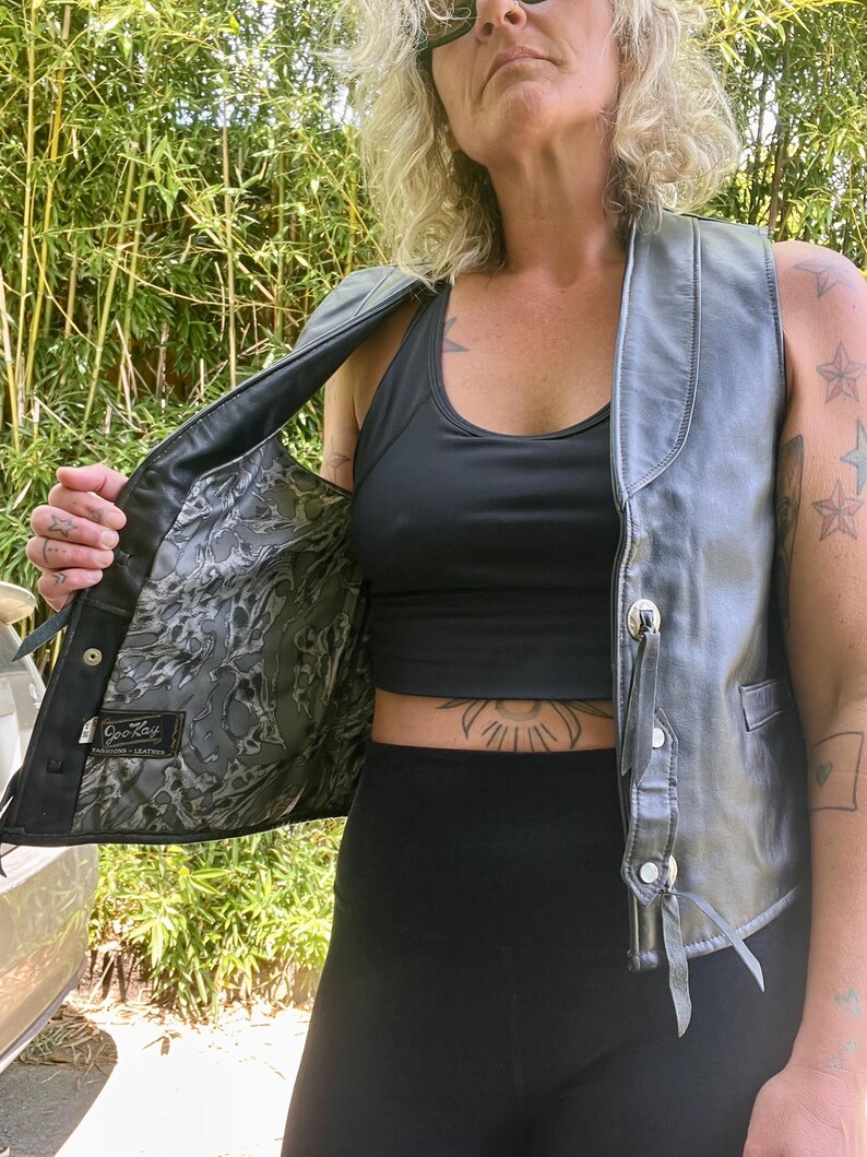 May include: A black leather vest with silver-toned buttons and decorative leather ties. The vest's interior features a patterned lining. The vest is open, revealing a black top and black leggings. The vest is a classic piece of apparel.
