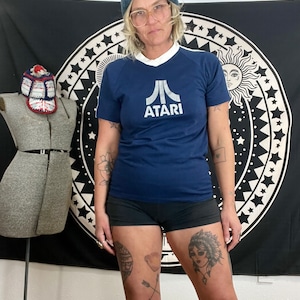 May include: A navy blue t-shirt with a white Atari logo and a white collar. The shirt has white stripes on the sleeves. The person wearing the shirt has a blue beanie and glasses.