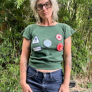 May include: A green top with patches, including a red cross and the words "Cookie Power." The top is worn with dark blue denim shorts. The background features green bamboo. The top has short sleeves.