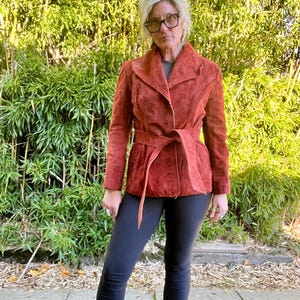 Könnte beinhalten: Eine rostbraune Wildlederjacke mit einem breiten Kragen, einem Bindegürtel und langen Ärmeln. Die Jacke wird über einer schwarzen Leggings getragen.