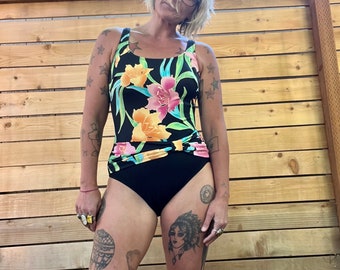 Pode incluir: Um maiô preto com estampa floral em tons de amarelo, rosa e verde. O maiô tem um decote redondo e costas baixas. A modelo está usando o maiô e tem tatuagens nos braços e nas pernas.