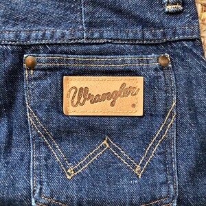 May include: A close-up of a blue denim pocket with a brown leather patch that says "Wrangler" in a script font. The pocket has a distinctive W-shaped stitching.