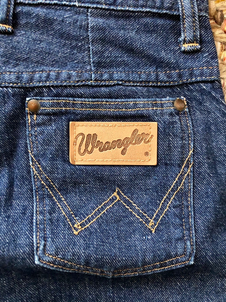May include: A close-up of a blue denim pocket with a brown leather patch that says "Wrangler" in a script font. The pocket has a distinctive W-shaped stitching.