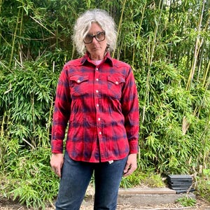 May include: A red and navy plaid button-down shirt with two chest pockets. The shirt is paired with dark wash blue jeans. The person is wearing glasses and standing in front of a bamboo background.