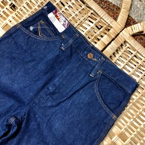 May include: Dark blue denim jeans with a brown leather patch and the text "Wrangler" on the back pocket. The jeans have a straight leg and a classic five-pocket style.