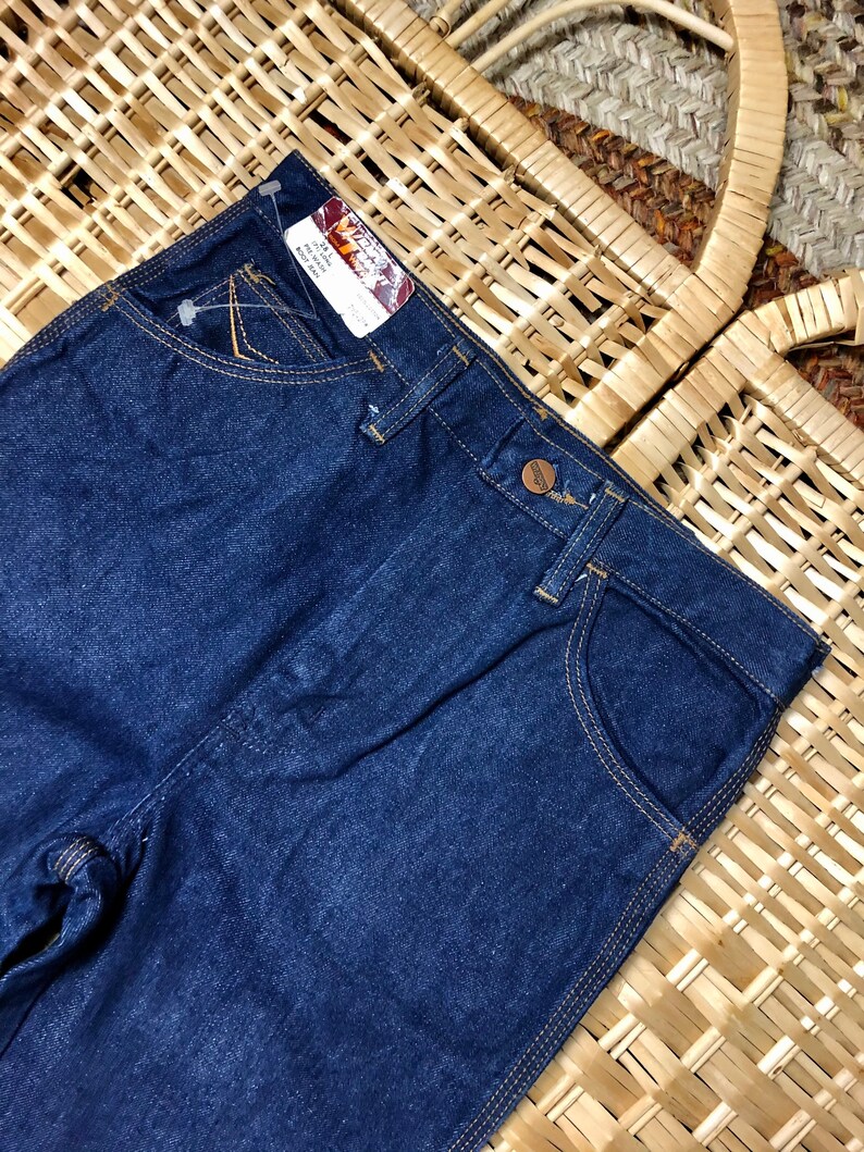 May include: Dark blue denim jeans with a brown leather patch and the text "Wrangler" on the back pocket. The jeans have a straight leg and a classic five-pocket style.