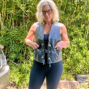 May include: A black leather vest with silver-colored buttons and pockets. The vest has a V-neck design and a tie closure. The vest is worn over a black top and paired with black leggings. The background features green bamboo.