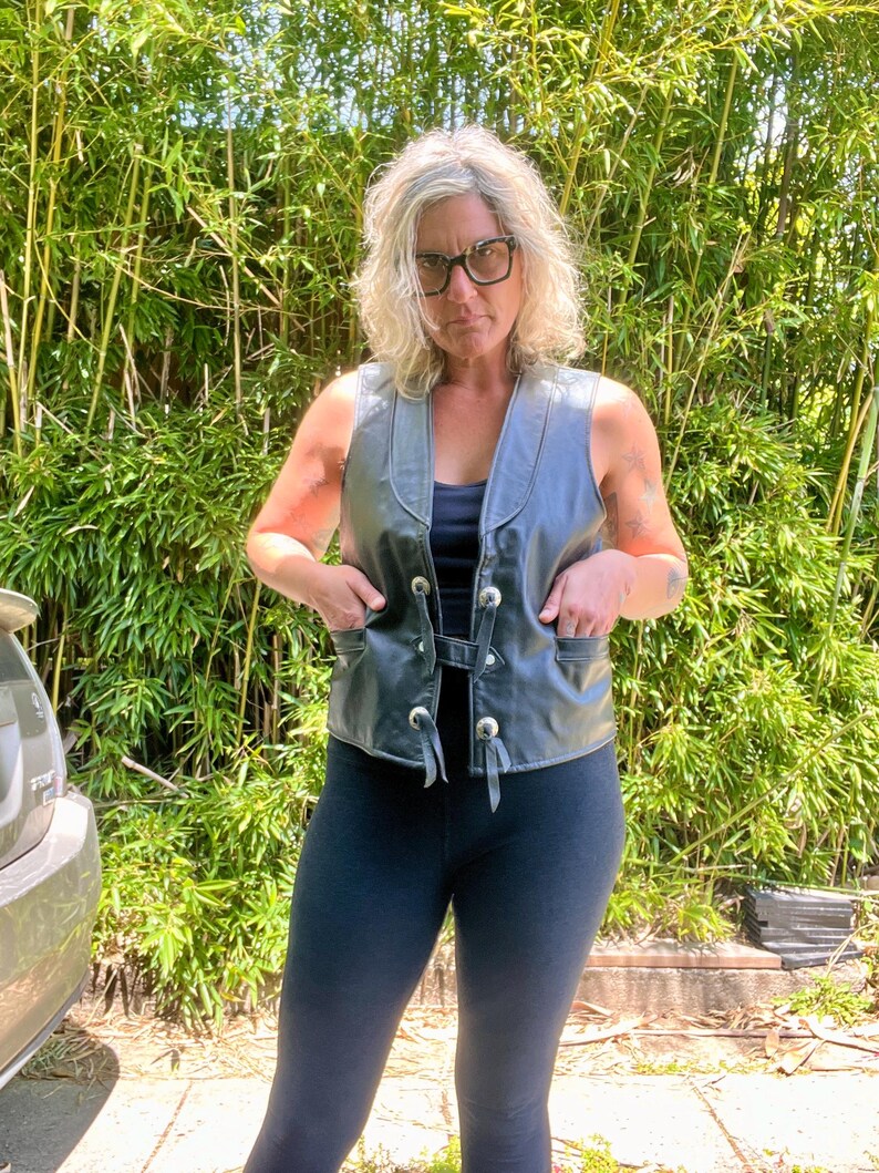 May include: A black leather vest with silver-colored buttons and pockets. The vest has a V-neck design and a tie closure. The vest is worn over a black top and paired with black leggings. The background features green bamboo.