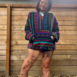 May include: A person wearing a colourful striped hooded sweater with a blue beanie and glasses. The sweater has a large pocket on the front. The person has tattoos on their legs.