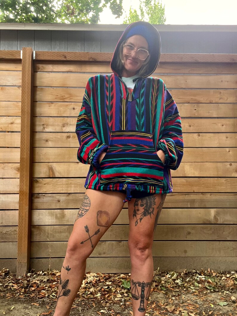 May include: A person wearing a colourful striped hooded sweater with a blue beanie and glasses. The sweater has a large pocket on the front. The person has tattoos on their legs.
