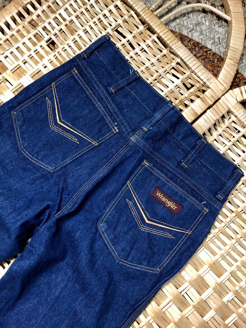 May include: A pair of dark blue denim jeans with a brown leather Wrangler label on the back pocket. The jeans have a classic five-pocket style and are made of a durable cotton blend.