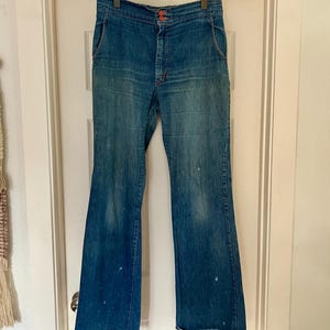 Może przedstawiać: Wzorzyste niebieskie dżinsy z dzwonami, z wysokim stanem i pomarańczowymi szwami. Dżinsy mają zapięcie na guziki i kieszenie. Denim ma wyblakły wygląd z kilkoma białymi plamami farby. Dżinsy wiszą na drewnianym wieszaku.