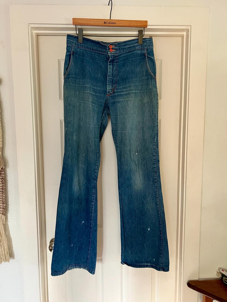 Może przedstawiać: Wzorzyste niebieskie dżinsy z dzwonami, z wysokim stanem i pomarańczowymi szwami. Dżinsy mają zapięcie na guziki i kieszenie. Denim ma wyblakły wygląd z kilkoma białymi plamami farby. Dżinsy wiszą na drewnianym wieszaku.