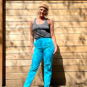 May include: A person wearing a brown beanie, grey tank top, and bright blue pants. The person is standing in front of a wooden fence.