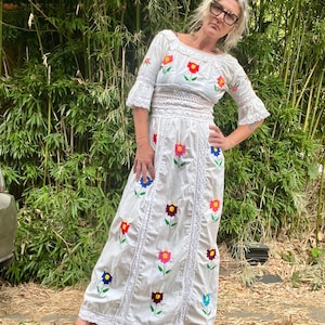 May include: A white, floor-length dress with colorful embroidered flowers. The dress features a scoop neckline, short sleeves, and lace trim. The flowers are red, yellow, blue, and purple. The person is wearing brown leather sandals.