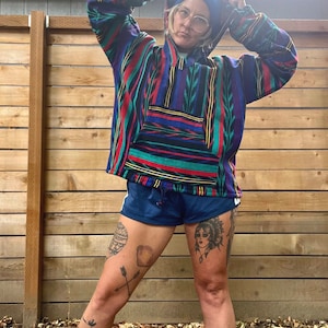 May include: A person wearing a colourful striped hooded sweatshirt with a blue beanie, blue shorts, and multiple tattoos on their legs. The person is standing in front of a wooden fence.