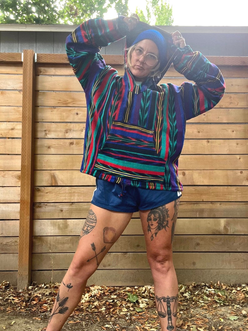 May include: A person wearing a colourful striped hooded sweatshirt with a blue beanie, blue shorts, and multiple tattoos on their legs. The person is standing in front of a wooden fence.