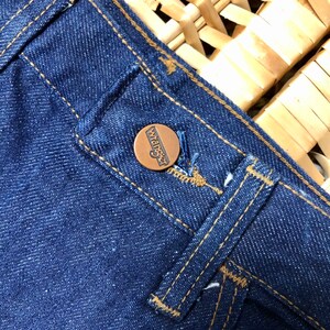 May include: Close-up of a blue denim jean pocket with a brown leather button that says "Wrangler" in white lettering.
