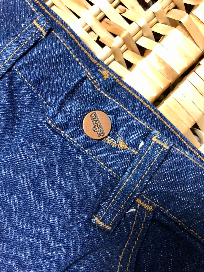 May include: Close-up of a blue denim jean pocket with a brown leather button that says "Wrangler" in white lettering.