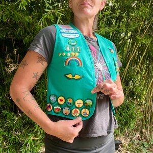 May include: A teal Girl Scout vest with various patches, including a rainbow, wings, and several circular badges. The vest has the number "477" and the words "Totem Council" on it. The vest is worn over a gray t-shirt.