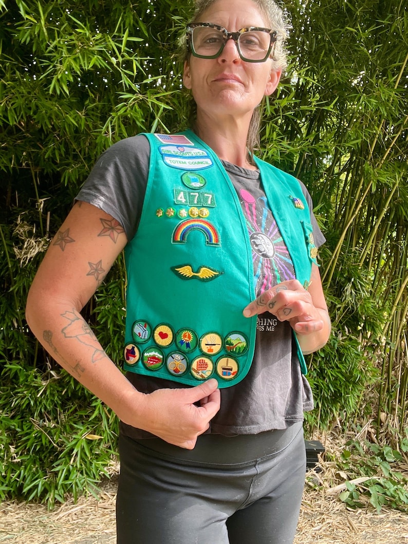 May include: A teal Girl Scout vest with various patches, including a rainbow, wings, and several circular badges. The vest has the number "477" and the words "Totem Council" on it. The vest is worn over a gray t-shirt.