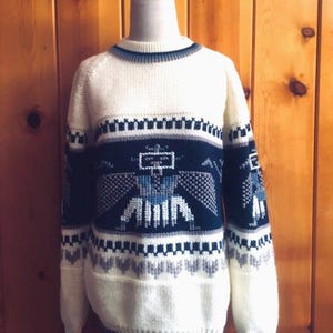 May include: A white knitted sweater with a blue, gray, and black geometric pattern. The pattern features a stylized bird design.