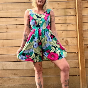 May include: A sleeveless floral print dress with a green and white pom-pom beanie. The dress has a fitted bodice and a flowing skirt. The model is wearing black high-top sneakers.