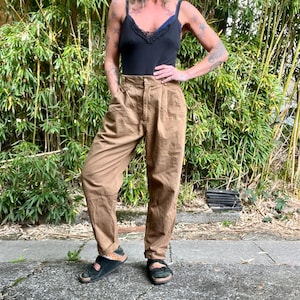 May include: A woman wearing a black camisole with lace trim and high-waisted brown pants. The pants have a pleated front and a relaxed fit. She is wearing black sandals with a patterned strap. The background features lush green bamboo.