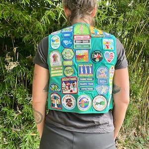 May include: A teal Girl Scout vest covered in colorful patches. The patches feature various designs, including birds, camping scenes, and text such as "Holiday Magic" and "Girl Scouts." The vest is worn over a gray t-shirt.