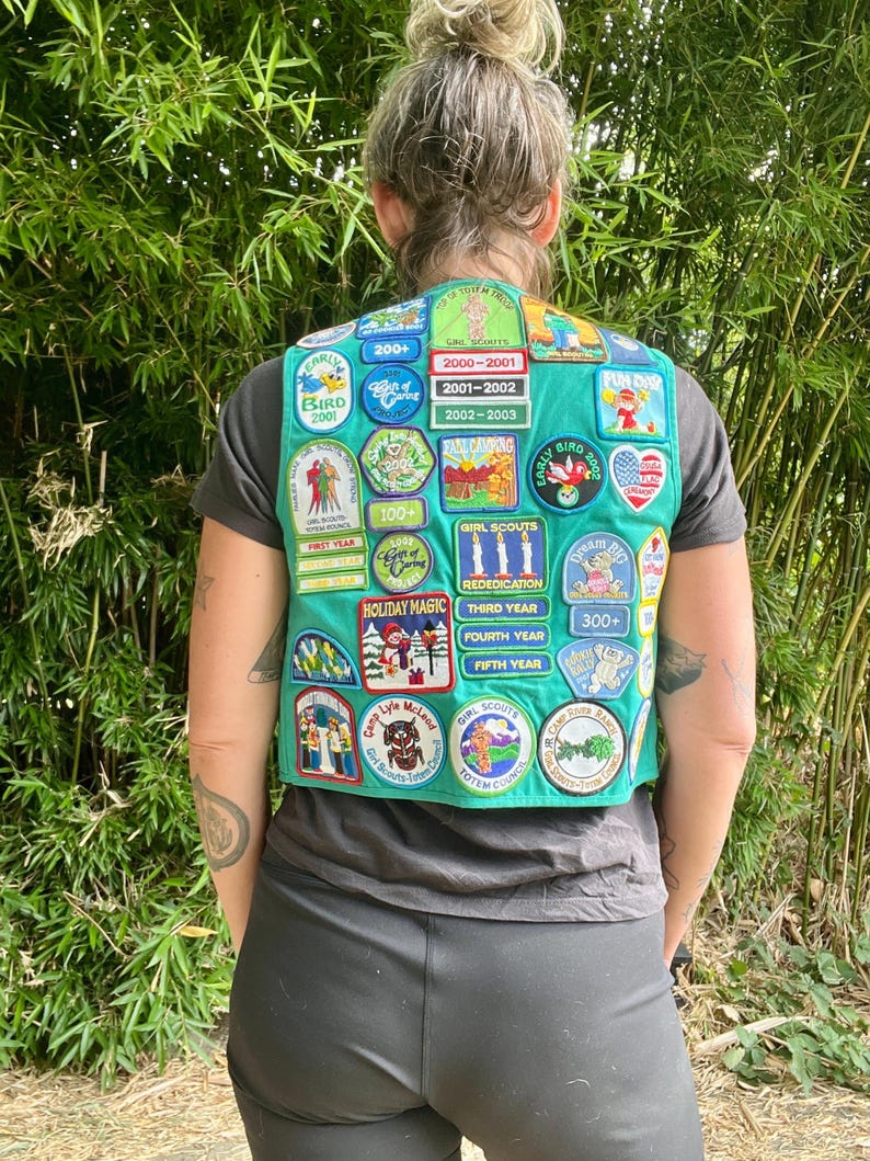 May include: A teal Girl Scout vest covered in colorful patches. The patches feature various designs, including birds, camping scenes, and text such as "Holiday Magic" and "Girl Scouts." The vest is worn over a gray t-shirt.