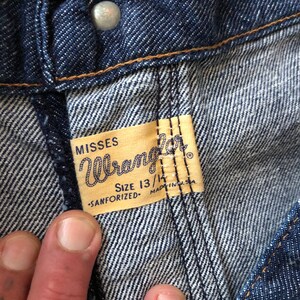 May include: Close-up of a blue denim jean label with the text "Misses Wrangler" and "Size 13/14 -Sanforized- Made in U.S.A."