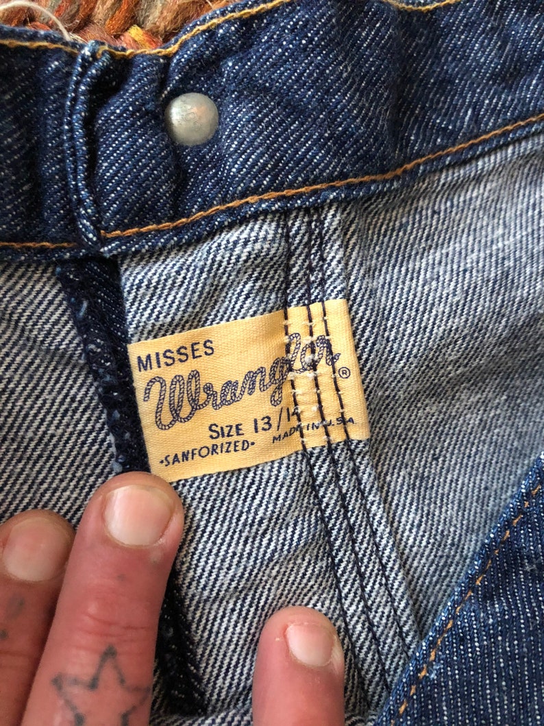 May include: Close-up of a blue denim jean label with the text "Misses Wrangler" and "Size 13/14 -Sanforized- Made in U.S.A."