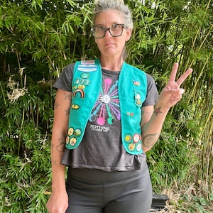 May include: A teal Girl Scout vest adorned with patches and pins, worn over a gray t-shirt with the text "Nothing Phases Me." The vest features a rainbow patch and various achievement badges. The person is wearing black shorts and glasses.