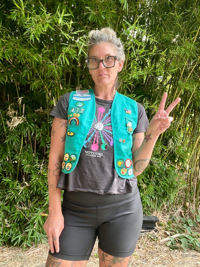May include: A teal Girl Scout vest adorned with patches and pins, worn over a gray t-shirt with the text "Nothing Phases Me." The vest features a rainbow patch and various achievement badges. The person is wearing black shorts and glasses.