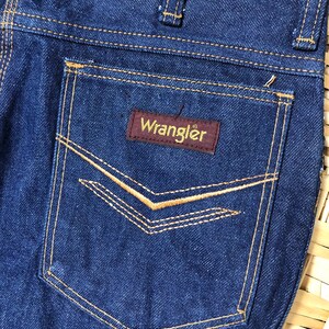 May include: A pair of dark blue denim jeans with a back pocket featuring a brown leather patch with the word "Wrangler" in yellow lettering.