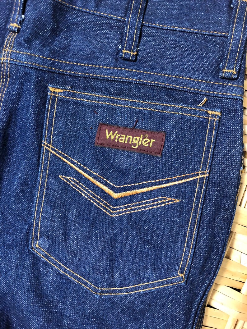 May include: A pair of dark blue denim jeans with a back pocket featuring a brown leather patch with the word "Wrangler" in yellow lettering.