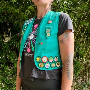 May include: Teal vest adorned with various patches and pins. The vest is worn over a black t-shirt with a colorful graphic and the text "NOTHING PHASES ME." The person is wearing black leggings.