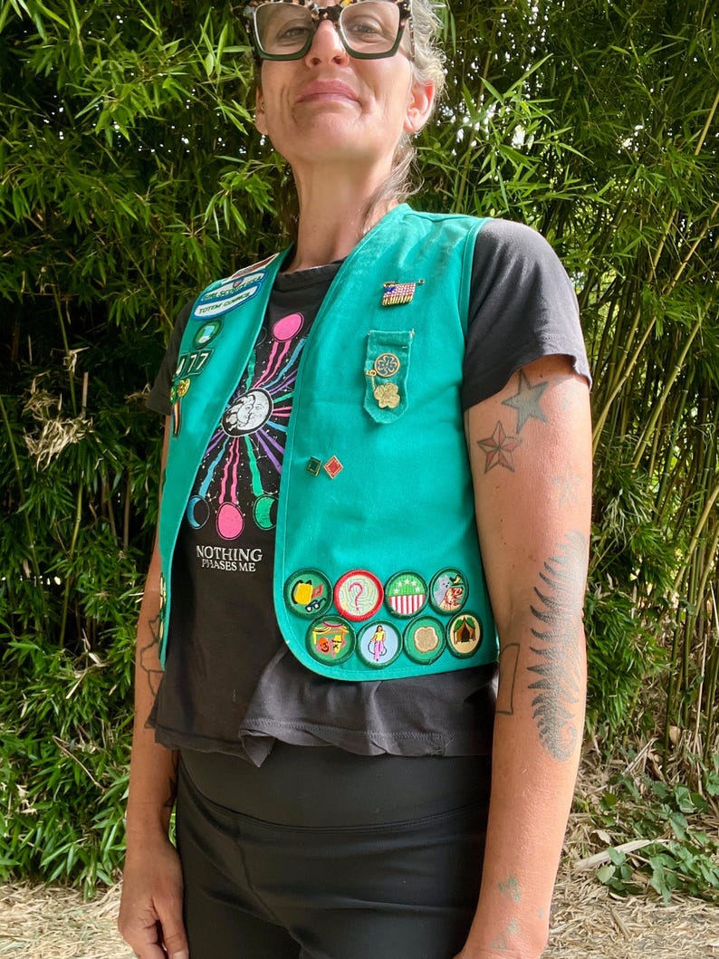 May include: Teal vest adorned with various patches and pins. The vest is worn over a black t-shirt with a colorful graphic and the text "NOTHING PHASES ME." The person is wearing black leggings.