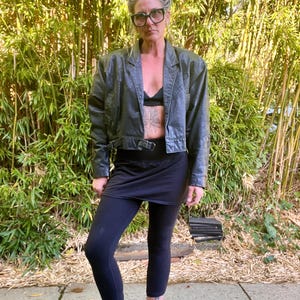 May include: A black leather jacket with a cropped design and a belt at the waist. The jacket has a classic collar and shoulder pads. The person is wearing a black bra, black skirt, and black leggings. They are also wearing black boots with pink laces.