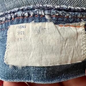 Może przedstawiać: Zbliżenie na dżinsową odzież z białą metką. Na metce widnieje napis "MENS SIZE 38 X 32". Dżins jest w kolorze jasnoniebieskim z pomarańczowymi przeszyciami. Metka ma wyblakłe logo.