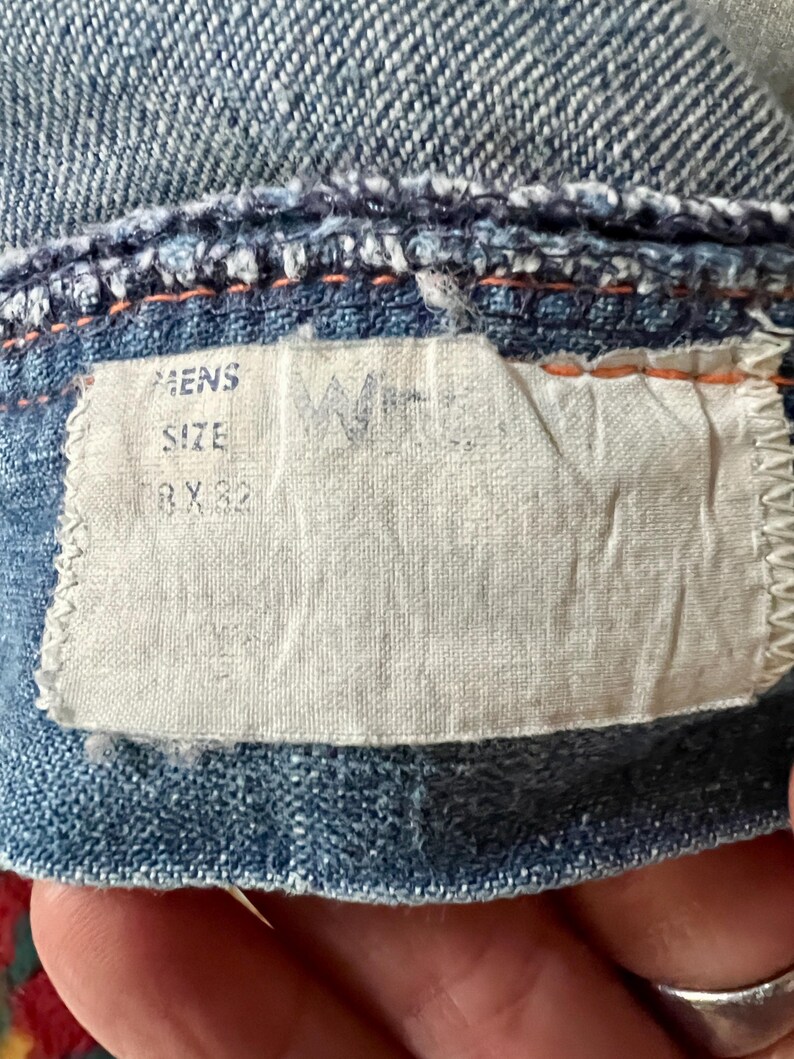 Może przedstawiać: Zbliżenie na dżinsową odzież z białą metką. Na metce widnieje napis "MENS SIZE 38 X 32". Dżins jest w kolorze jasnoniebieskim z pomarańczowymi przeszyciami. Metka ma wyblakłe logo.