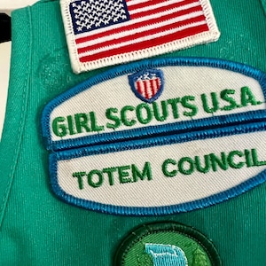 May include: Close-up of a green Girl Scout uniform with embroidered patches. The patches include an American flag, "Girl Scouts USA," and "Totem Council." A circular patch features a waterfall scene. The patches are in various colors.