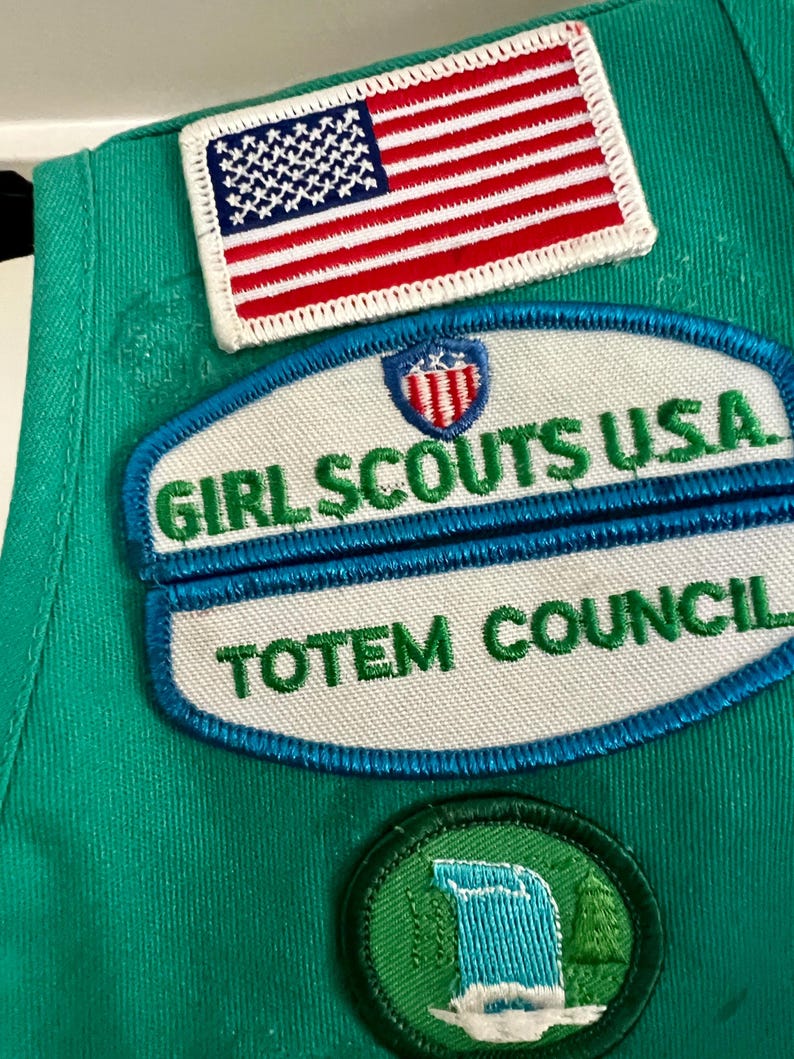 May include: Close-up of a green Girl Scout uniform with embroidered patches. The patches include an American flag, "Girl Scouts USA," and "Totem Council." A circular patch features a waterfall scene. The patches are in various colors.
