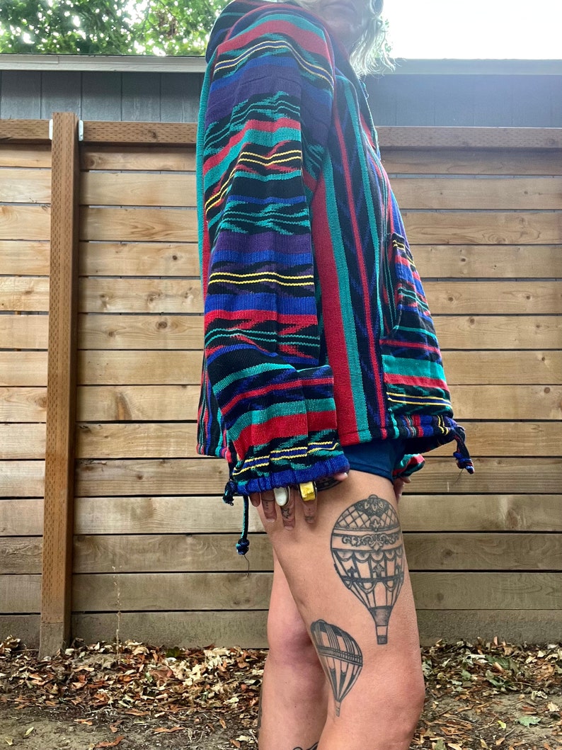 May include: A colourful striped jacket with a zip closure. The jacket is made of a soft, woven fabric. The person wearing the jacket has a tattoo of hot air balloons on their leg.
