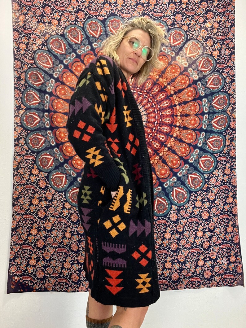 May include: A black long-sleeved cardigan with a geometric pattern in shades of orange, yellow, purple, and green. The cardigan has a shawl collar and a relaxed fit.