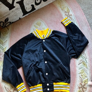 May include: A black satin bomber jacket with yellow and white striped trim around the collar, cuffs, and hem. The jacket has a button closure and two pockets.