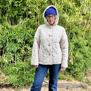 May include: A cream-colored quilted jacket with a hood and toggle closures. The jacket is worn by a person wearing a blue beanie and blue jeans.
